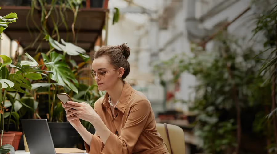 A young woman in glasses and a brown shirt uses her phone while sitting in a lush, plant-filled indoor space.