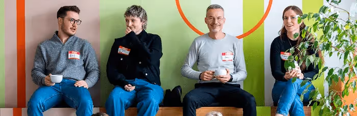 Four people sit against a colorful wall, drinking coffee and engaged in conversation