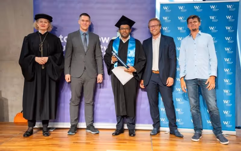 Graduate in cap and gown poses with university faculty and leaders during a graduation ceremony.