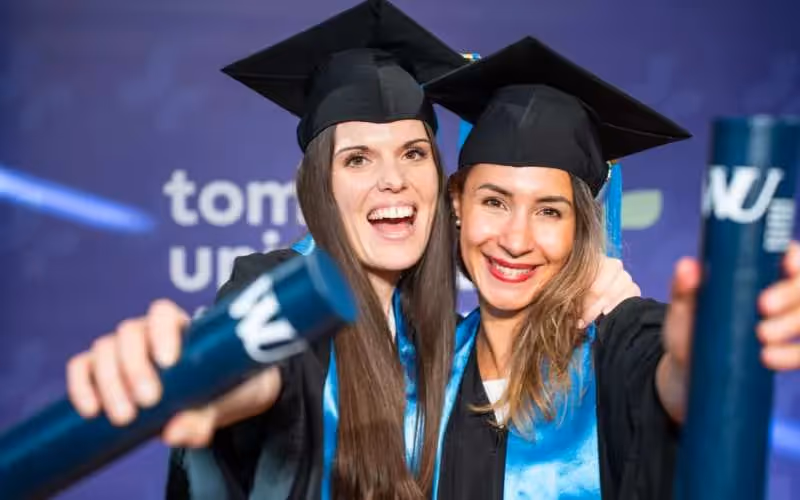 Two joyful graduates in caps and gowns celebrate, holding diplomas and smiling at the camera.