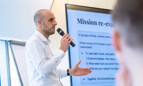Man presenting with a microphone in front of a screen titled "Mission re-evaluation