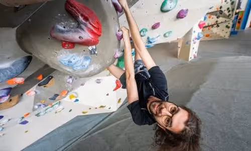 Smiling man climbing indoors on a colorful bouldering wall, hanging upside down.