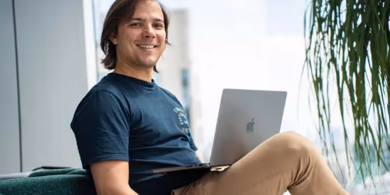 Henrique smiling while using a laptop, sitting by a window with a green background.