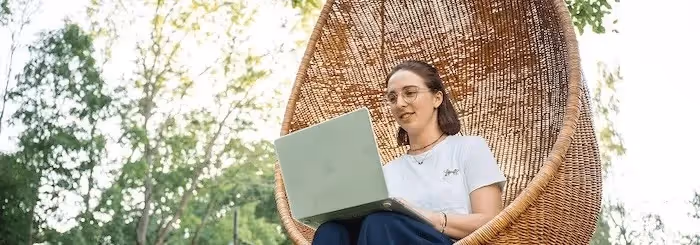 A woman sits in a hanging wicker chair outdoors, working on a laptop with a smile