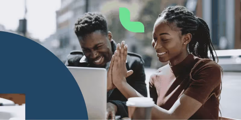 Zwei junge schwarze Menschen, die im Freien abklatschen und lächelnd gemeinsam an einem Laptop arbeiten.