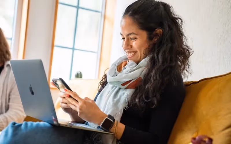 Smiling woman using smartphone and laptop while sitting on a yellow couch.