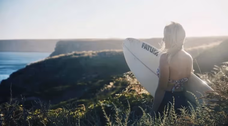 Bianca with surfboard gazes at ocean cliffs under soft morning light.