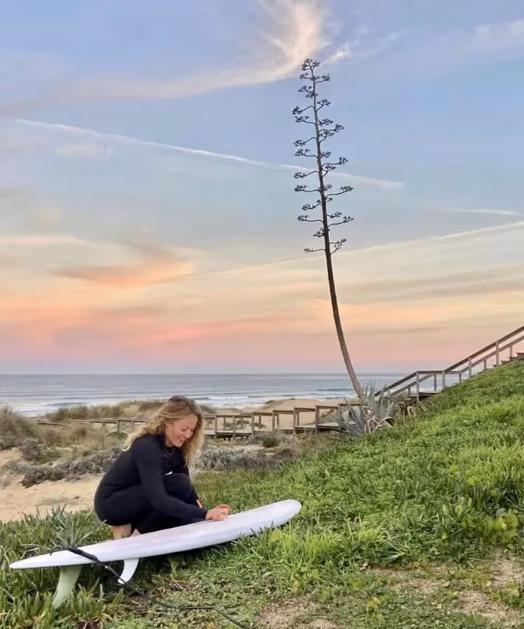 Bianca kneels to wax surfboard on grassy dune with pastel sunset and ocean behind.