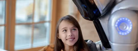 Young girl smiling at a humanoid robot in a warmly lit room.