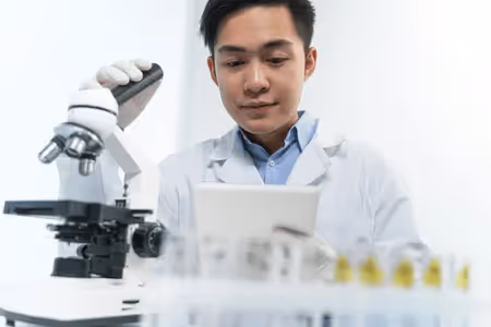 A researcher sitting in his lap and looking at a tablet, while touching his microscope.