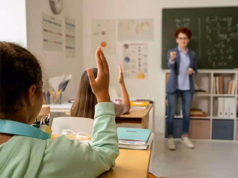 Children raising hands in a classroom while teacher points toward them