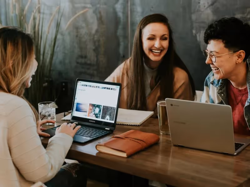 Three students laughing and working on laptops at a wooden table