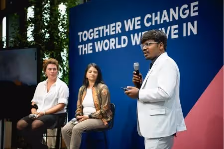 Man speaking at Green Tech Festival panel with slogan backdrop and two seated panelists