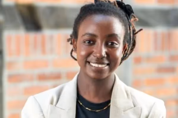 Vinley Orutwa, lächelnd in einem hellen Blazer vor einer Backsteinwand.