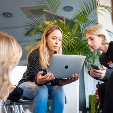 A woman with light brown hair looks at a laptop while discussing with two other women in an office space.