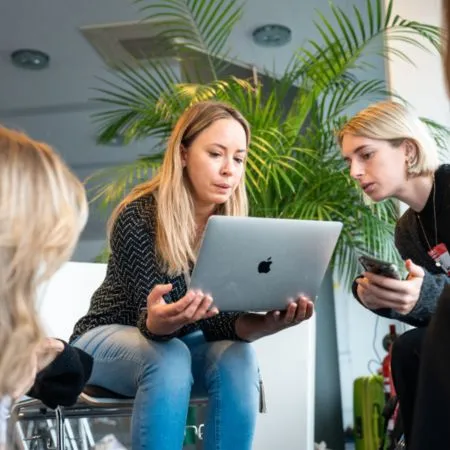 A woman with light brown hair looks at a laptop while discussing with two other women in an office space.