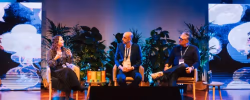 Three people sit on stage in discussion with plants and large screens behind them, engaged in a conference or panel event.