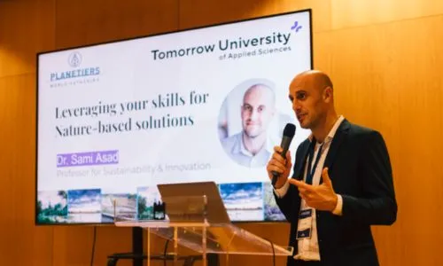 A man speaks at a podium with a microphone, next to a large screen showing his photo and the title "Leveraging your skills for Nature-based solutions."