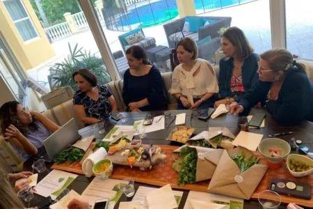 Group of women sitting around a table full of food and herbs, engaged in a lively discussion and tasting session.
