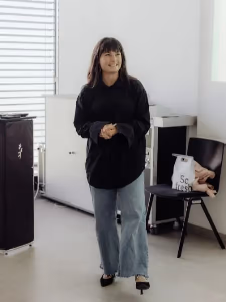 Woman standing in a bright room wearing a black shirt and jeans, smiling softly, with a chair and personal items in the background.