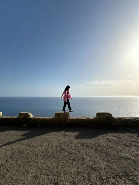 Silhouette of a person walking on a narrow stone wall with a vast blue ocean and clear sky in the background.