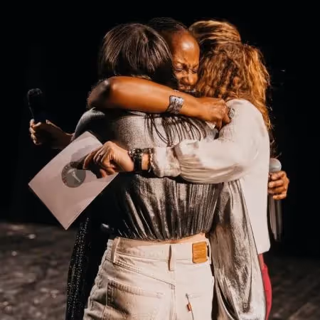 Three women embrace tightly on stage, two holding microphones and one holding a paper, sharing an emotional moment of connection and support.