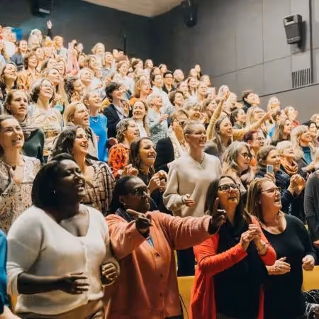 Large group of diverse women smiling and cheering together in an auditorium, celebrating and sharing a joyful moment.