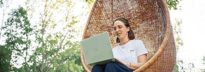 Eine Frau sitzt in einem hängenden Weidenkorbstuhl im Freien und arbeitet lächelnd an einem Laptop.