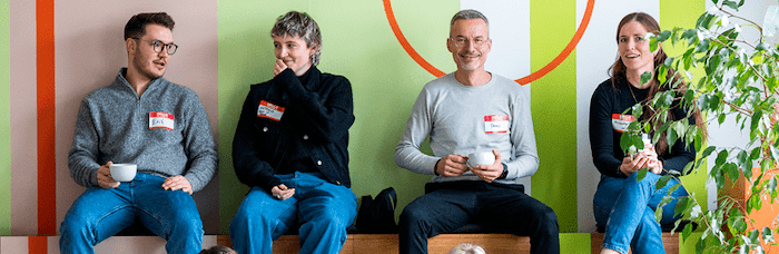 Vier Personen sitzen vor einer bunten Wand, trinken Kaffee und führen ein Gespräch.