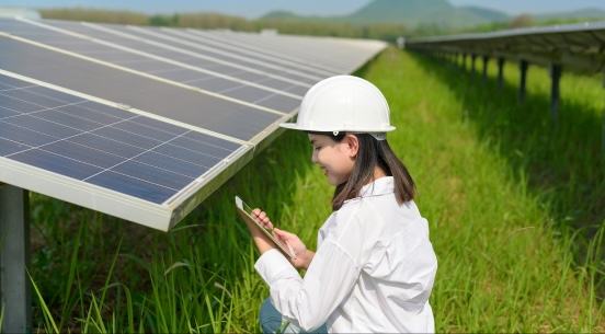Frau mit Schutzhelm inspiziert Solarpanele mit einem Smartphone in einem grasbewachsenen Feld.