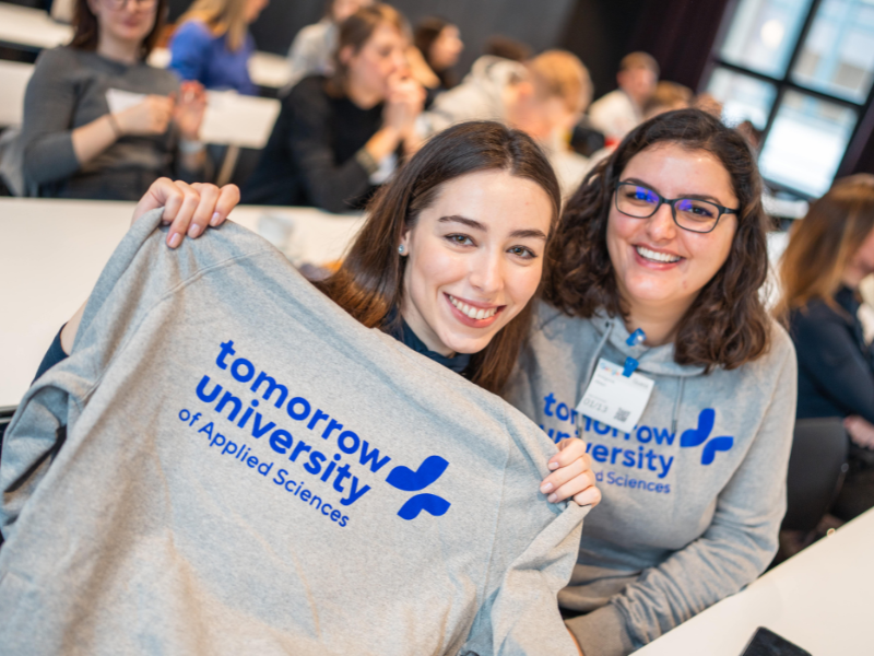 Zwei Frauen lächeln und halten einen Tomorrow University Hoodie bei einem Community-Treffen im Klassenzimmer.