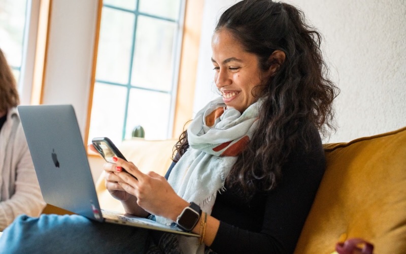 Lächelnde Frau, die Smartphone und Laptop benutzt, während sie auf einem gelben Sofa sitzt.