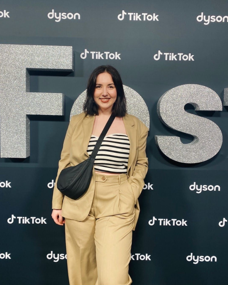 Frau im beigefarbenen Anzug und gestreiftem Oberteil posiert vor einer TikTok- und Dyson-Eventkulisse.