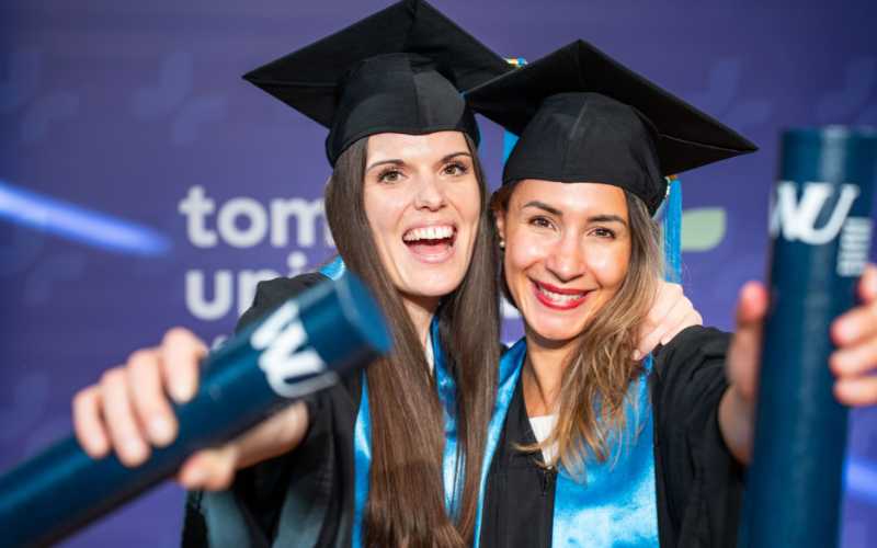 Zwei freudige Absolventen im Talar feiern, halten Diplome und lächeln in die Kamera.