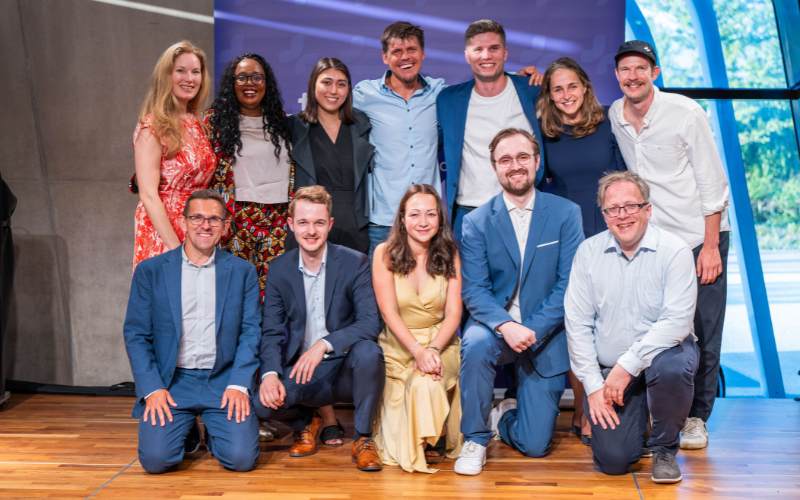 Gruppenfoto von Dozenten und Mitarbeitern, die zusammen auf einer Holzbühne bei der Abschlussfeier lächeln.