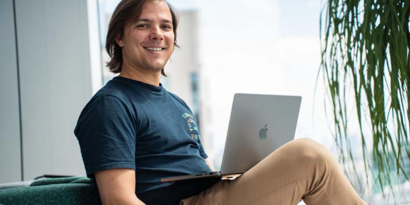 Henrique lächelt beim Arbeiten an einem Laptop, sitzt neben einem Fenster mit grünem Hintergrund.