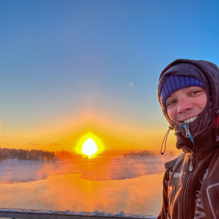 Henrique in Winterkleidung lächelnd vor einem leuchtend orangefarbenen Sonnenaufgang über einer schneebedeckten, eisigen Landschaft.