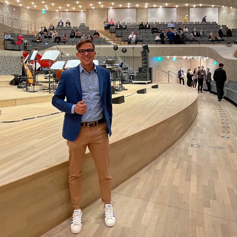 Henrique in einem blauen Blazer in einem Konzertsaal nahe einer Bühne mit Instrumenten und sitzenden Menschen.