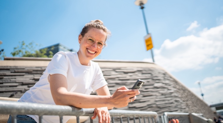 Marie mit Brille lächelt, während sie ein Telefon hält und sich an ein Geländer lehnt, mit Himmel und Laternenpfahl darüber.
