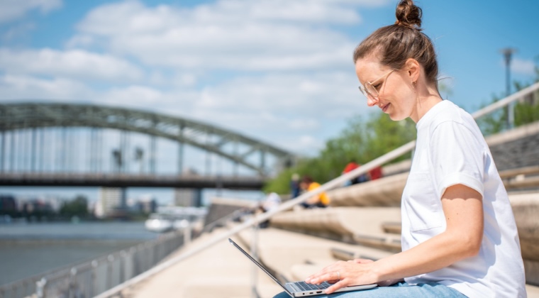 Marie im weißen Hemd arbeitet an einem Laptop, während sie auf einer Ufertreppe nahe einer großen Brücke sitzt.