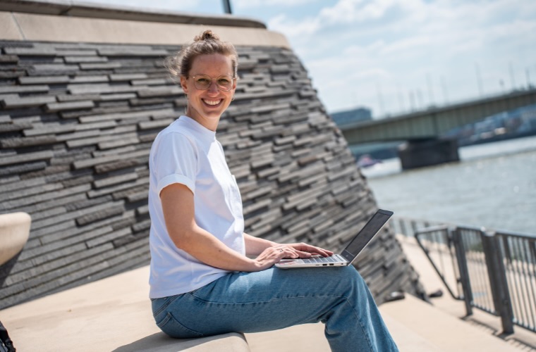 Marie tippt auf einem Laptop, während sie an einer Ufermauer sitzt, mit einer Brücke im Hintergrund.