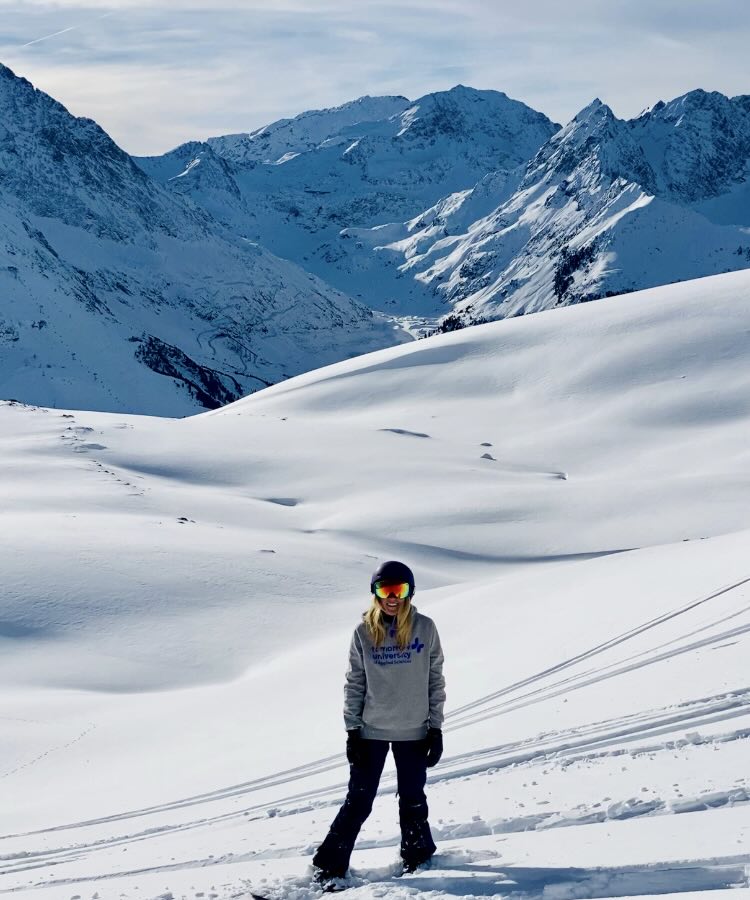 Bianca in Skiausrüstung steht auf verschneiter Piste mit Berggipfeln im Hintergrund.