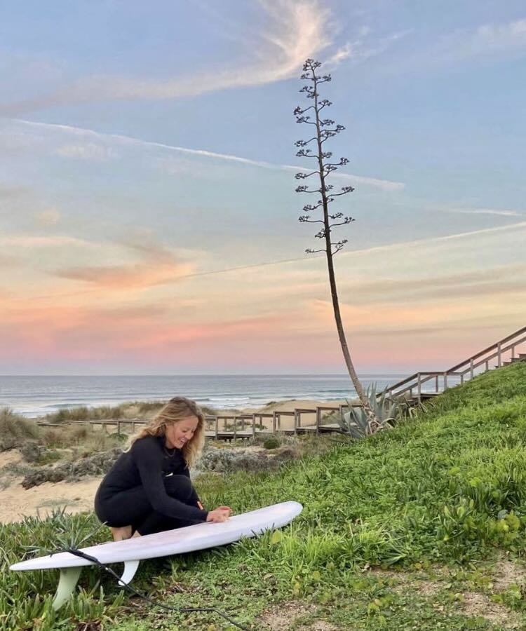 Bianca kniet, um ein Surfbrett auf einer grasbewachsenen Düne zu wachsen, mit pastellfarbenem Sonnenuntergang und Ozean im Hintergrund.