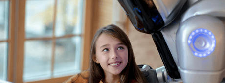 Junges Mädchen lächelt einen humanoiden Roboter in einem warm beleuchteten Raum an.