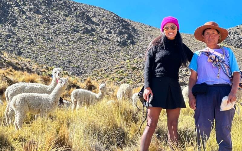 Frau in Schwarz mit rosa Hut und einem Einheimischen in einem Feld mit Alpakas im Hintergrund.
