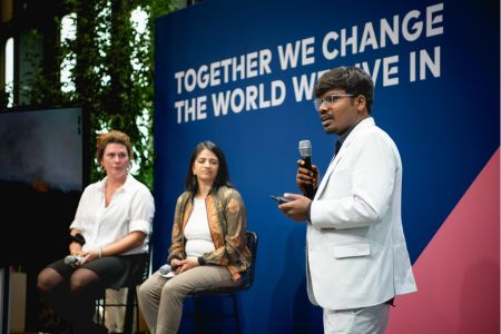 Mann spricht auf einer Green Tech Festival Podiumsdiskussion mit Slogan-Hintergrund und zwei sitzenden Panelist:innen