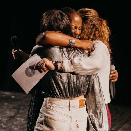 Drei Frauen umarmen sich fest auf der Bühne, zwei halten Mikrofone und eine hält ein Papier, teilen einen emotionalen Moment der Verbindung und Unterstützung.