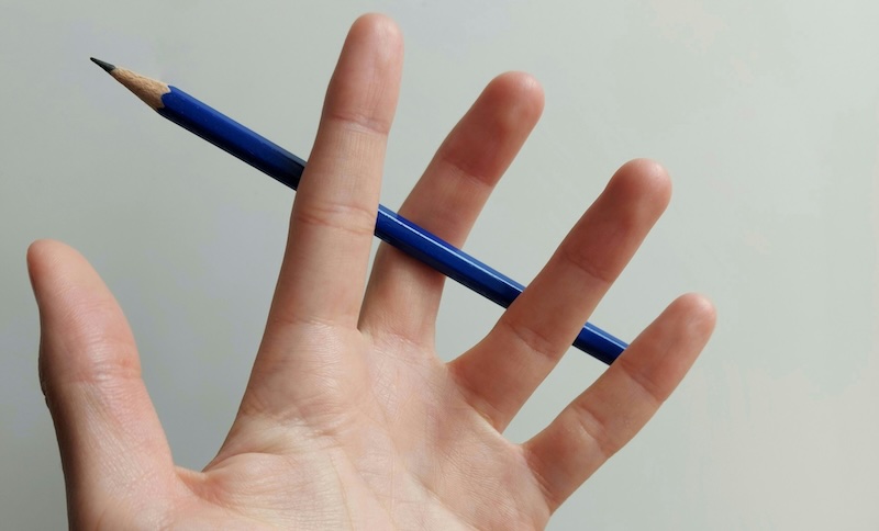 A hand holding a blue pencil loosely between its fingers against a plain light background.