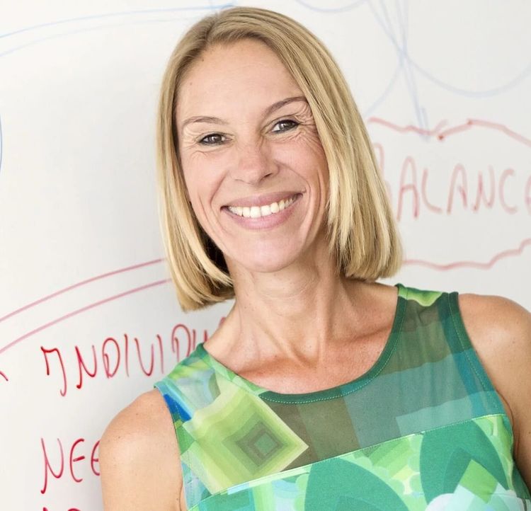 Smiling woman in a green patterned sleeveless top standing in front of a whiteboard with handwritten notes.