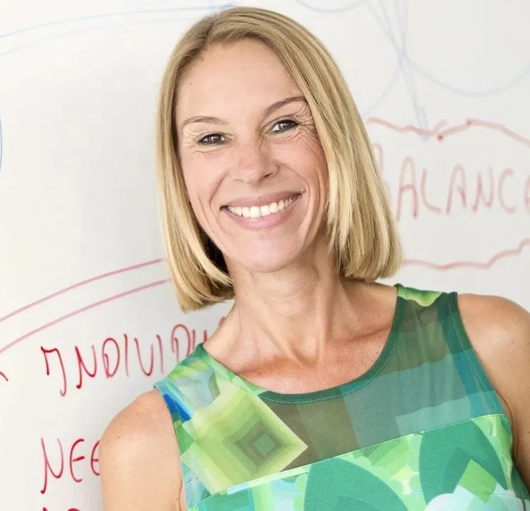 Smiling woman in a green patterned sleeveless top standing in front of a whiteboard with handwritten notes.
